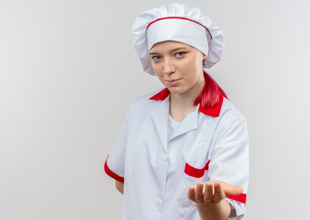 De jonge tevreden blonde vrouwelijke chef-kok in eenvormige chef-kok houdt uitdelen geïsoleerd op witte muur