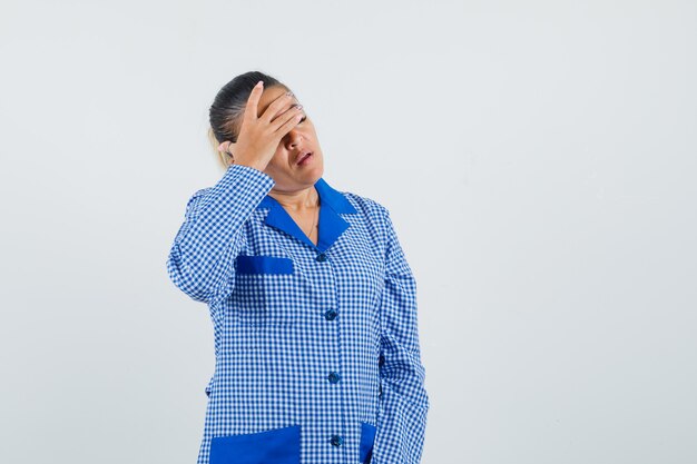 De jonge vrouw die een deel van het gezicht behandelt met dient het blauwe overhemd van de gingangpyjama in en ziet er moe uit. vooraanzicht.