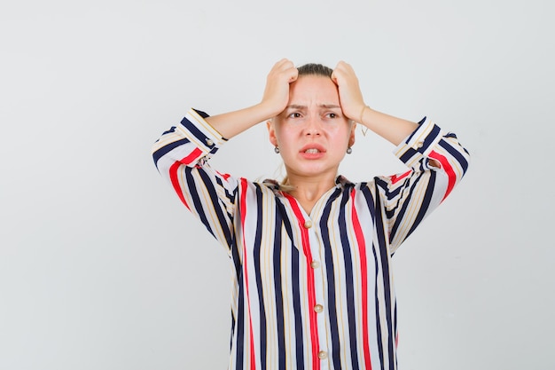 Gratis foto de jonge vrouw die haar hoofd behandelt met beide dient gestreepte blouse in en kijkt uitgeput