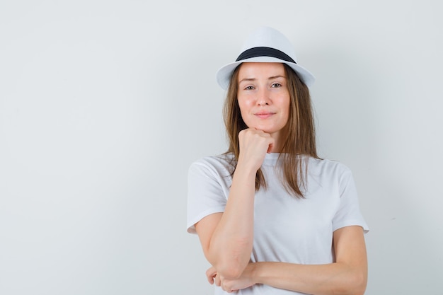 De jonge vrouw die kin aan hand steunt in wit t-shirt, hoed en op zoek zelfverzekerd.