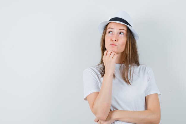 De jonge vrouw die kin op hand in wit t-shirt, hoed steunt en besluiteloos kijkt.