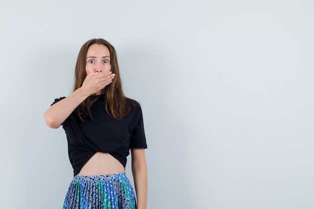 De jonge vrouw die mond behandelt dient zwarte t-shirt en blauwe rok in en kijkt verbaasd
