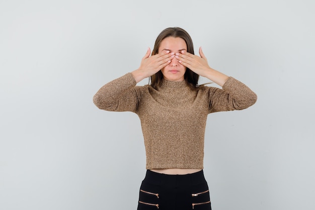 Gratis foto de jonge vrouw die ogen behandelt met handen in goud vergulde sweater en zwarte broek en ernstig, vooraanzicht kijkt.