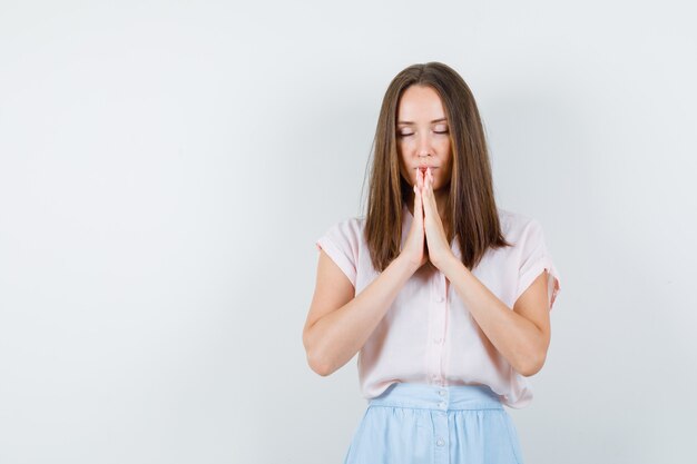 De jonge vrouw dient biddend gebaar in t-shirt, rok te houden en kalm te kijken. vooraanzicht.