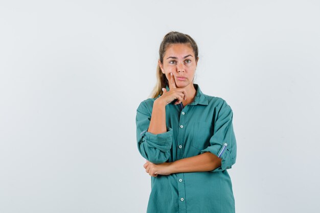 De jonge vrouw in groene blouse die zich in het denken stelt stelt en peinzend kijkt