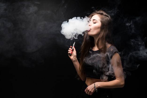 De jonge vrouw in zwarte rookt een elektronische sigaret op donkere muur
