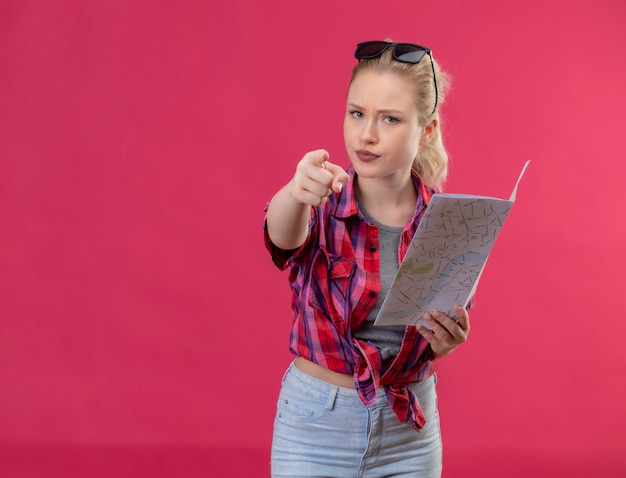 De jonge vrouwelijke reiziger die een rood overhemd en een bril op haar hoofd draagt met kaart toont je gebaar op geïsoleerde roze muur