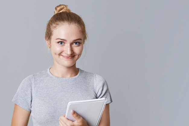 De jonge vrouwelijke student met spiraalvormig notitieboekje propt voor examen, herziet al materiaal, terloops gekleed, geïsoleerd op grijs met exemplaarruimte voor uw reclame. studeren concept