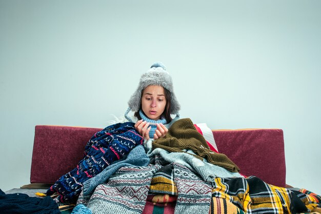 De jonge zieke vrouw met rookkanaal zittend op de Bank thuis of studio bedekt met warme gebreide kleding. Ziekte, griep, pijn concept. Ontspanning thuis. Gezondheidszorgconcepten.