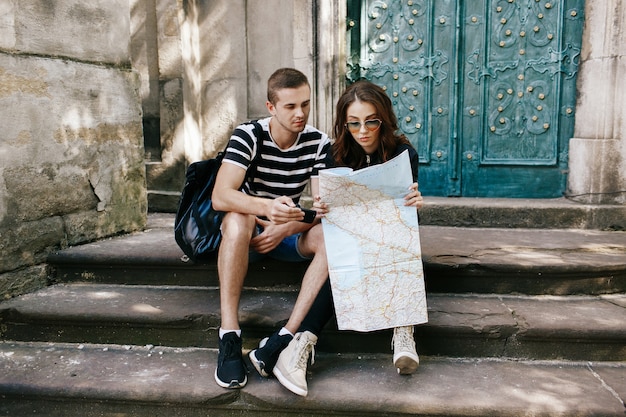 De jongen en het meisje zitten op de stappen aan kathedraal met touristckaart en letten op iets in smartphone