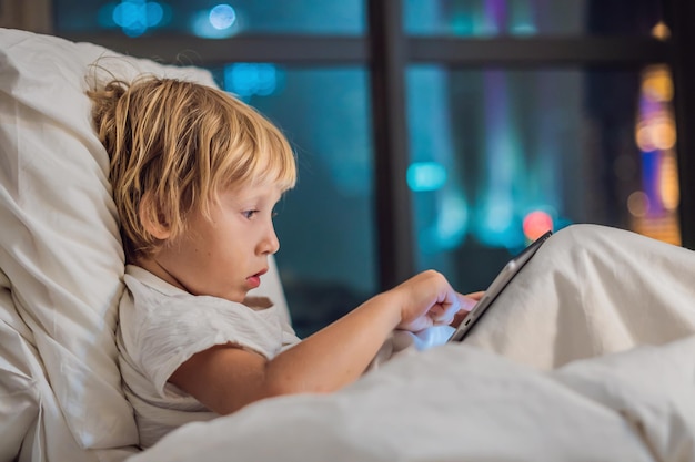 De jongen gebruikt de tablet in zijn bed voordat hij gaat slapen op een achtergrond van een nachtstad. kinderen en technologieconcepten