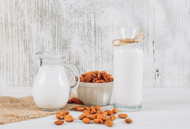 De karaf van de zijaanzichtmelk met kom van amandelen en fles melk op witte houten en stuk van zakachtergrond. horizontaal