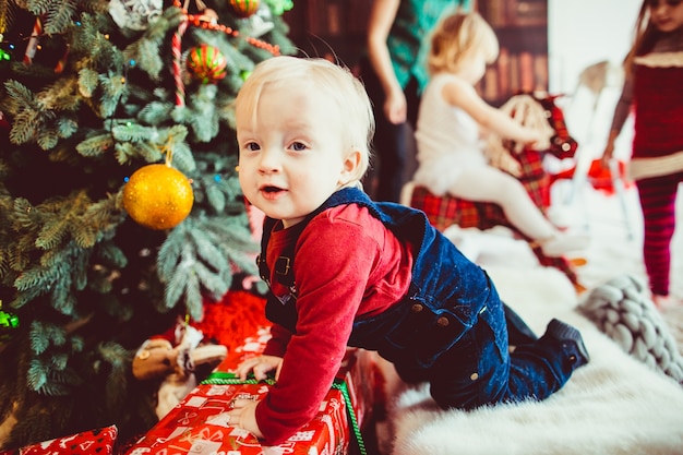 De kleine jongen zit in de buurt van de kerstboom en presenteert