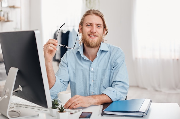 De knappe vrolijke gebaarde jonge blonde mannelijke copywriter typt informatie voor reclame op website, draagt blauw overhemd en bril, zit bij coworking kantoor voor het scherm.