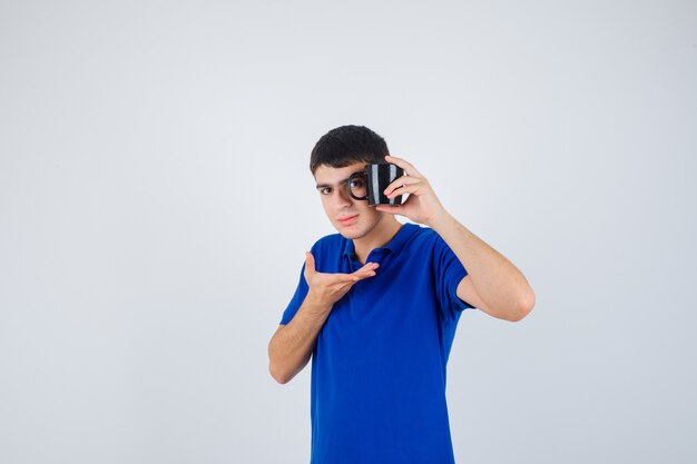 De kop van de jonge jongensholding, hand uitrekken als iets in blauw t-shirt vasthouden en ernstig, vooraanzicht kijken.