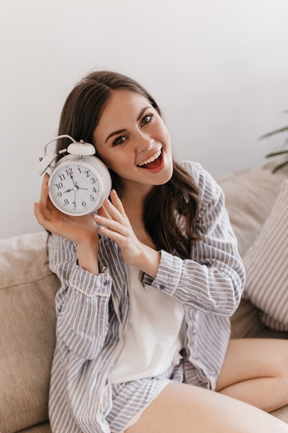 De leuke vrouw in mooie pyjama glimlacht, bekijkt voorzijde en houdt wekker vast