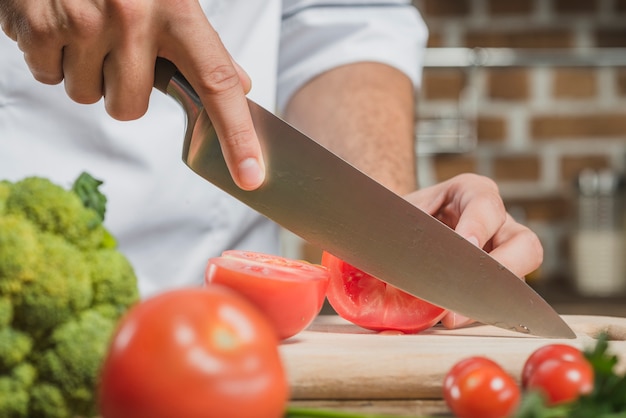 De mannelijke hand die van de chef-kok de tomaat met scherp mes aan boord snijdt