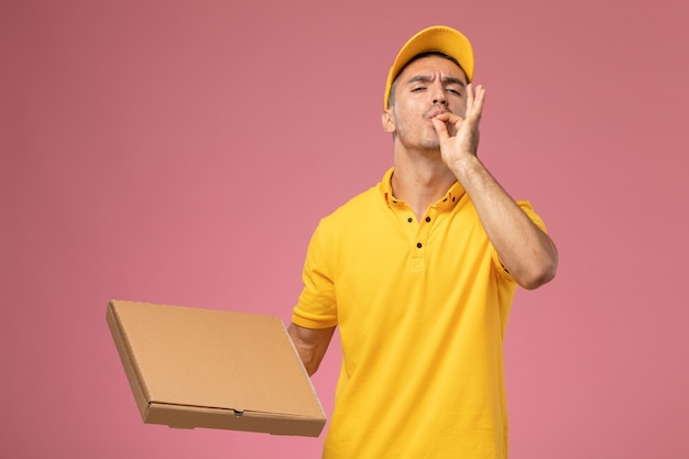 De mannelijke koerier van het vooraanzicht in de gele uniforme doos van de voedsellevering toont het smakelijke teken op roze bureau