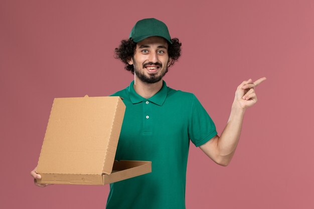 De mannelijke koerier van het vooraanzicht in groen uniform en cape die een lege doos van het leveringsvoedsel op roze achtergronddienstbaan uniforme levering houdt