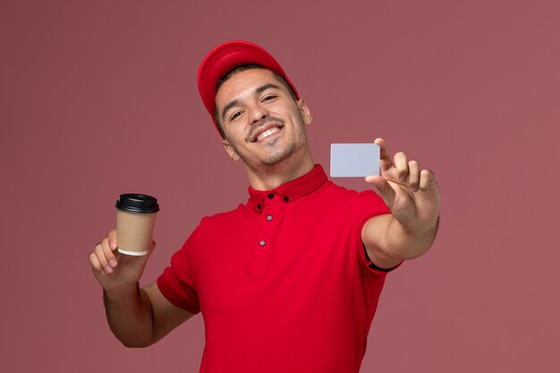 De mannelijke koerier van het vooraanzicht in rood uniform de koffiekop en de kaart van de holdingslevering op de roze bureaumedewerker