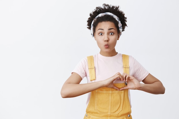 De mens heeft liefde nodig. portret van hoopvolle schattige en tedere afrikaanse amerikaan in gele overall en hoofdband, hartgebaar tonen in de buurt van borst en lippen vouwen in kus, poseren over grijze muur