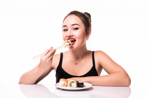 De mooie Aziaat kijkt met rode lippen zit op de lijst eet sushi rolt glimlachen geïsoleerd op wit