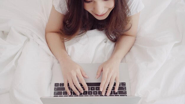 De mooie Aziatische Kaukasische vrouw die van het mengelingsras computer of laptop met behulp van terwijl het liggen op het bed in haar slaapkamer.