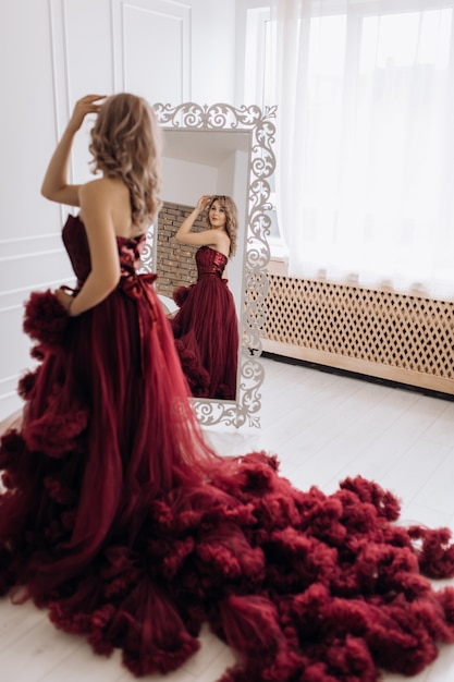 De mooie blondevrouw in kleding van luxe de rode Bourgondië stelt vóór een spiegel in een witte ruimte