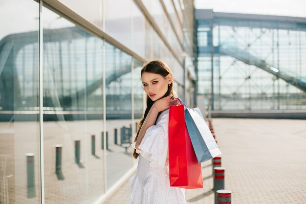 De mooie brunette met lang haar stelt met het winkelen zakken vóór een modern glasgebouw