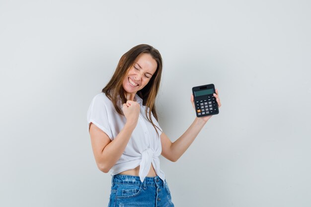 De mooie calculator van de dameholding terwijl het winnaargebaar in witte blouse, jeans wordt getoond en energiek eruit ziet. vooraanzicht.