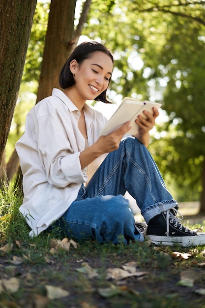 De mooie jonge Aziatische studente zit in park onder boom en leest boek glimlachend genietend van warme su