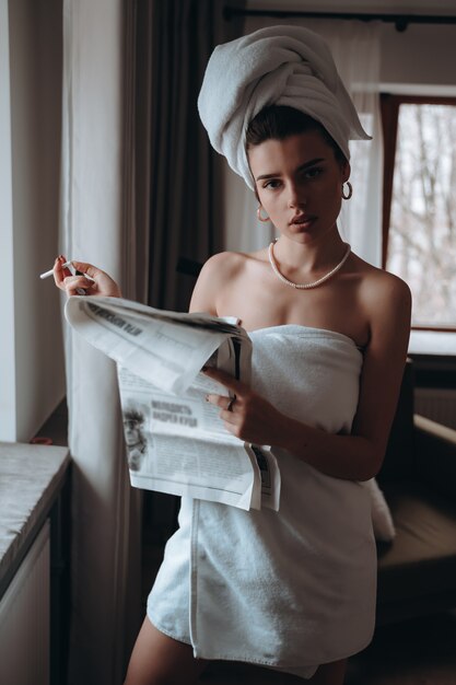 De mooie jonge vrouw in een handdoek rookt een sigaret en leest krant