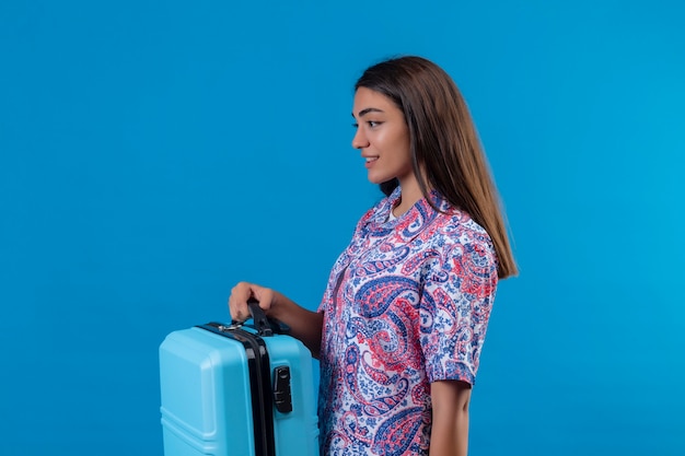 De mooie koffer die van de de holdingsreis van de reizigersvrouw zelfverzekerd met glimlach op gezicht kijkt die zich zijwaarts over geïsoleerde blauwe ruimte bevindt