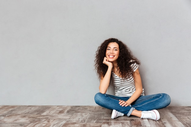 De mooie krullende vrouw die in vrijetijdskleding in lotusbloem zitten stelt op de vloer die haar hoofd met hand steunt die gelukkig en openhartig over grijze muur zijn