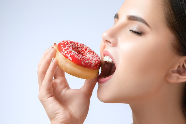 De mooie vrouw die een doughnut bijt bij grijze studioachtergrond