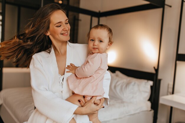 De mooie vrouw met glimlach bekijkt haar baby. Moeder in badjas die met baby in haar armen in slaapkamer dwarrelt.