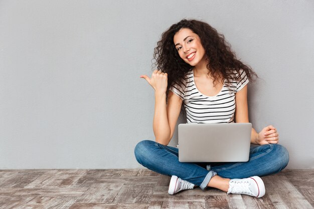 De mooie vrouw met mooie glimlachzitting in lotusbloem stelt op de vloer met zilveren computer op benen die opzij duim gesturing die iets exemplaarruimte indienen