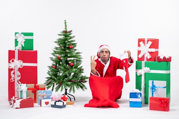 De nerveuze Kerstman toont twee zittend op de grond en houdt kerstsok in de buurt van geschenken en versierde nieuwe jaarboom op witte achtergrond