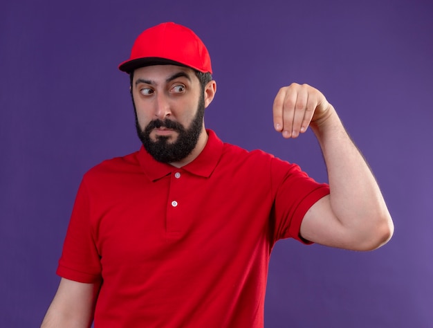 De onder de indruk jonge knappe blanke bezorger met een rood uniform en een pet doet alsof hij iets vasthoudt en naar zijn hand kijkt die op paars wordt geïsoleerd