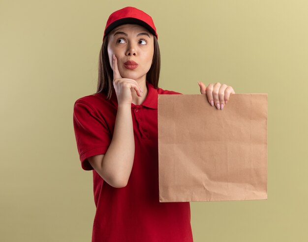 De onder de indruk mooie bezorgvrouw in uniform legt de vinger op het gezicht en houdt het papieren pakket vast, kijkend naar de zijkant op olijfgroen