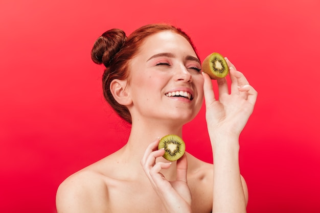 De opgewonden Europese kiwi van de meisjesholding. Studio shot van zorgeloze vrouw met exotische vruchten geïsoleerd op rode achtergrond.