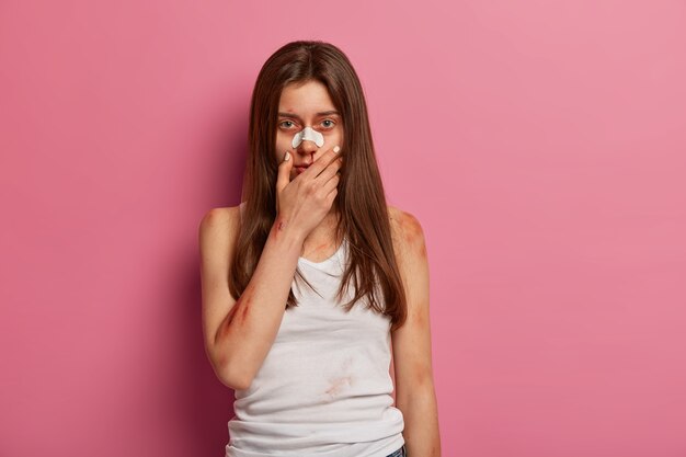 De overstuur vrouw kreeg een huisongeval vanwege onzorgvuldig gedrag. Gewonde vrouw slachtoffer van geweld houdt hand op gezicht, heeft gekneusde huid, bloedende wonden, poseert tegen roze muur. Verzekering.