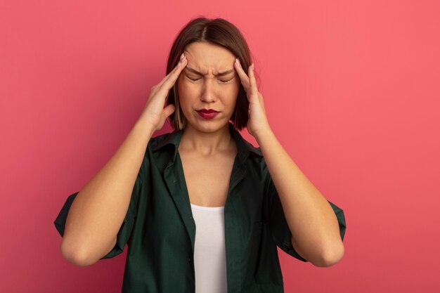 De pijnlijke mooie vrouw legt handen op tempels die op roze muur worden geïsoleerd