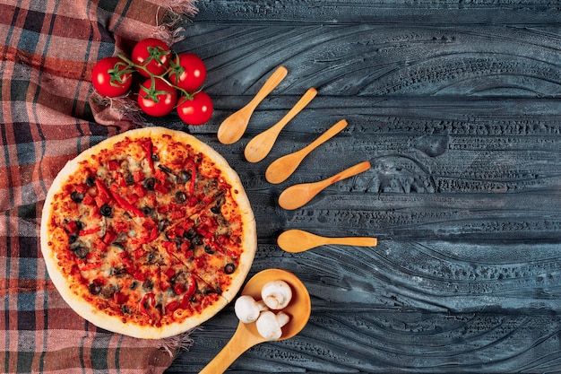 De pizza met tomaten, paddestoelen, houten lepelsvlakte lag op een donkere houten en picknickdoekachtergrond
