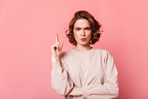 De prachtige gekrulde vrouw heeft een idee. Nadenkend Kaukasisch meisje dat met vinger op roze achtergrond richt.