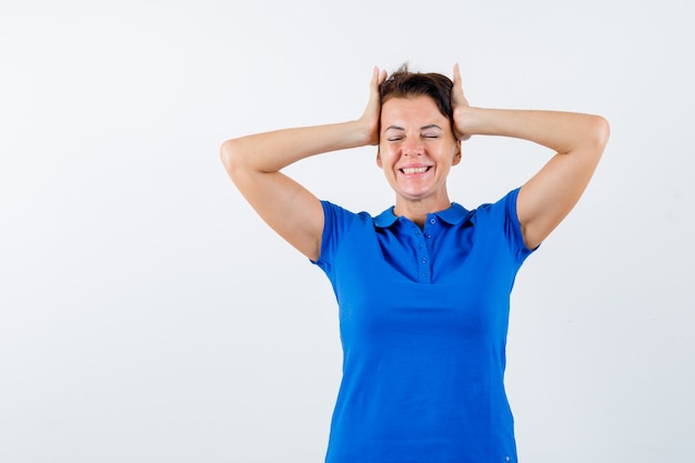 De rijpe vrouw die het hoofd met handen in blauw t-shirt vastklemt en zalig kijkt. vooraanzicht.