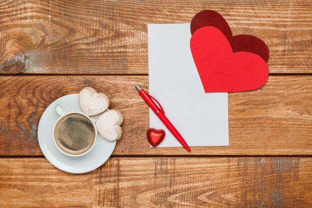 De rode harten en blanco vel papier en pen op houten achtergrond met een kopje koffie