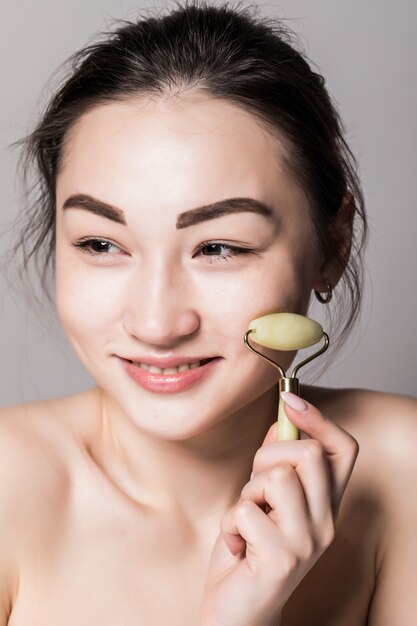 De schoonheid nam het gezichtsrol van de jadesteen voor gezichtsmassagetherapie toe die op grijze muur wordt geïsoleerd. Portret van Aziatische vrouw.