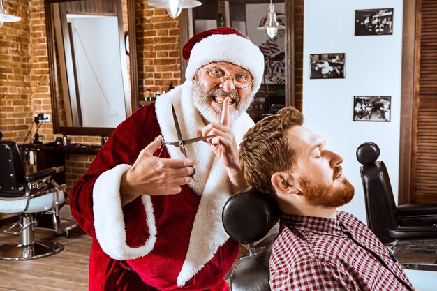 De senior man in kerstman kostuum werkt als persoonlijke meester met een schaar bij de kapper voor Kerstmis