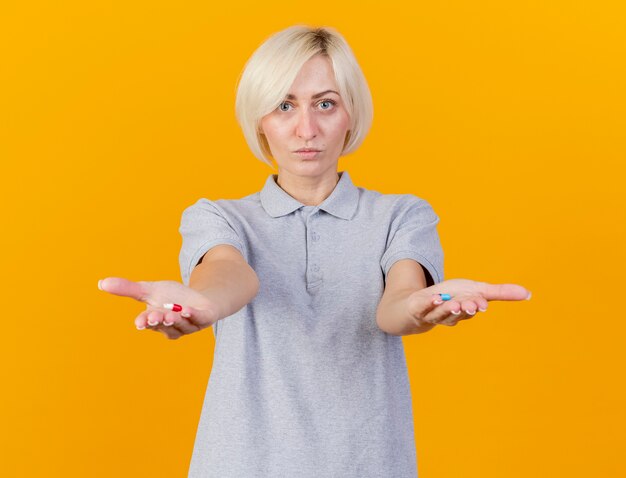 De tevreden jonge blonde zieke vrouw houdt medische pillen die op oranje muur worden geïsoleerd